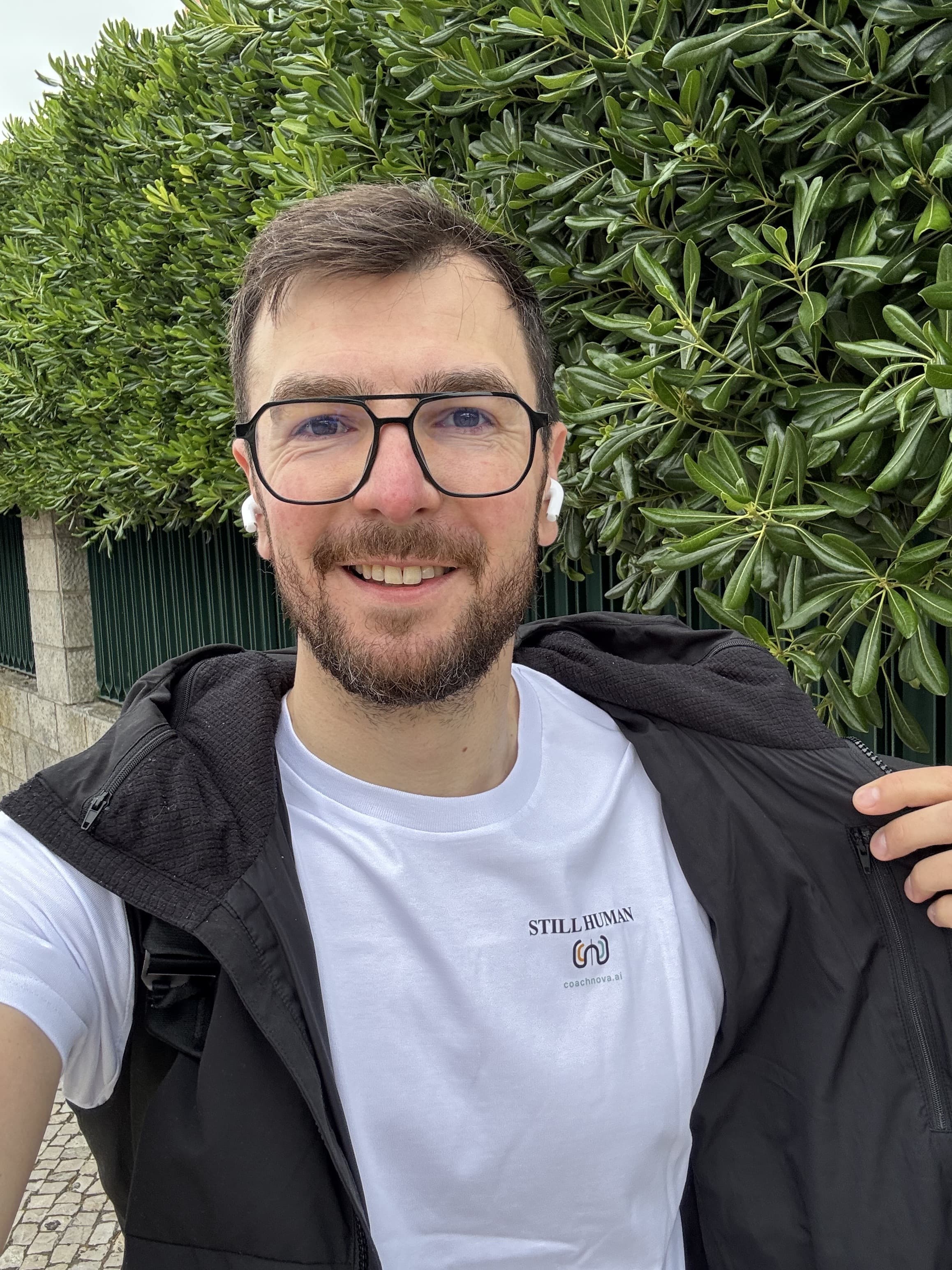 Nick wearing the white CoachNova Still Human t-shirt, with the Still Human wordmark and coachnova.ai logo, against a green hedge.