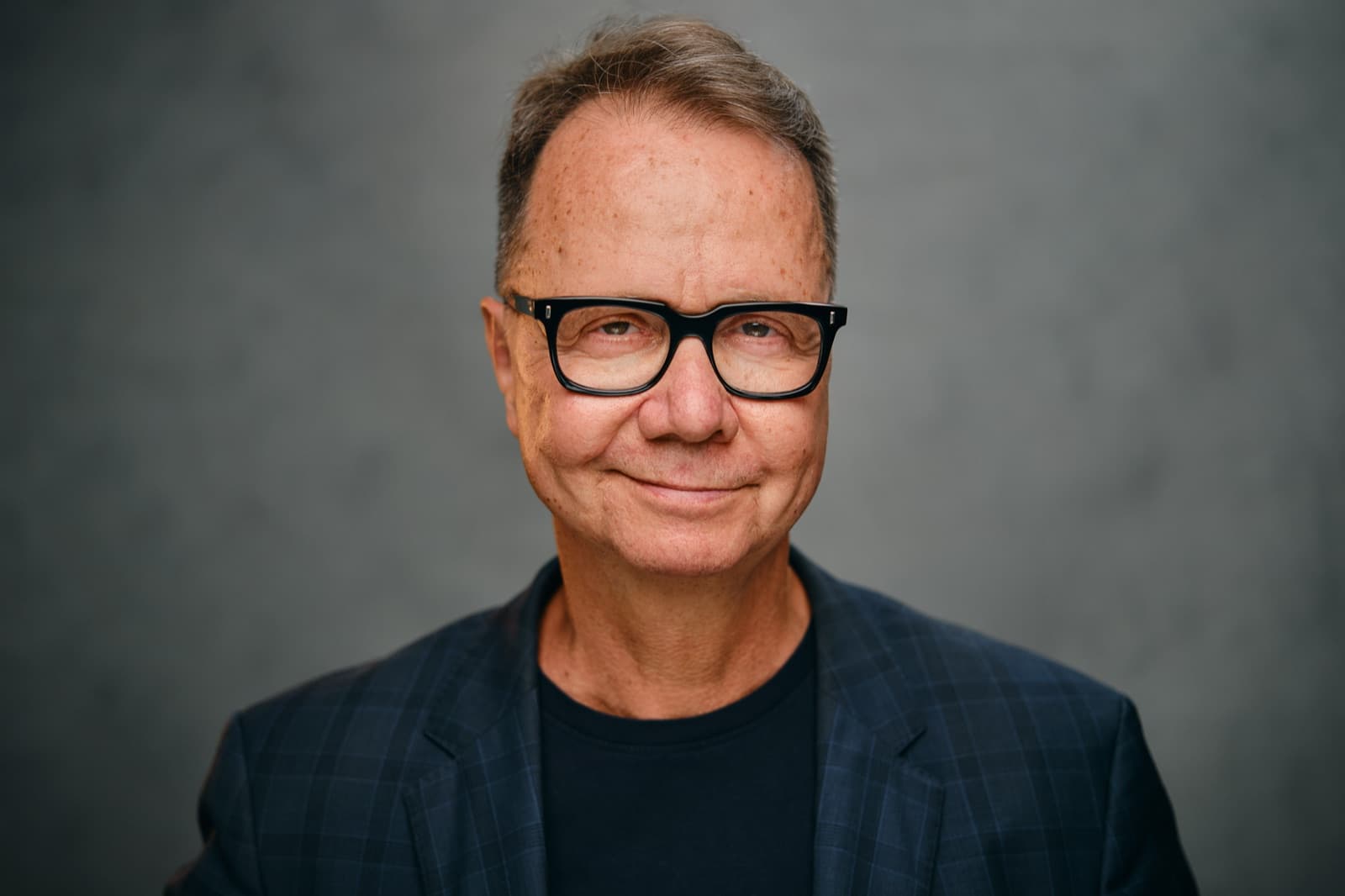Achim Nowak, smiling, in dark-rimmed glasses and a navy check blazer against a grey backdrop.