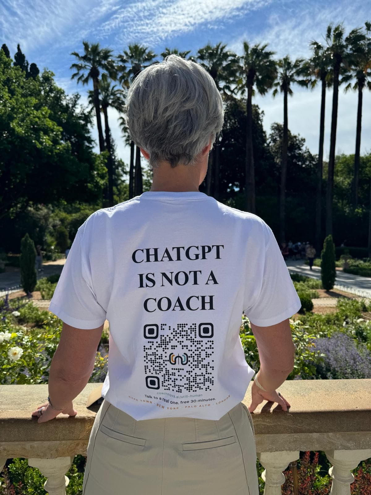 Louise from behind in a white Still Human t-shirt reading CHATGPT IS NOT A COACH, with a CoachNova QR code on the back, looking out at palm trees at the Zappeion gardens in Athens.